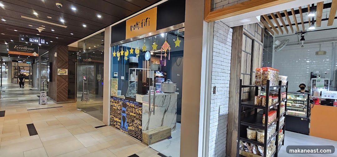 The exterior of a shop called Roti Fifi, with a bright orange sign above the entrance. The shop has a blue facade with yellow star decorations. Inside, a counter displays various food items and a drink dispenser. To the right, shelves are stocked with packaged snacks, and a display case holds cakes. A person wearing a mask stands behind the counter.