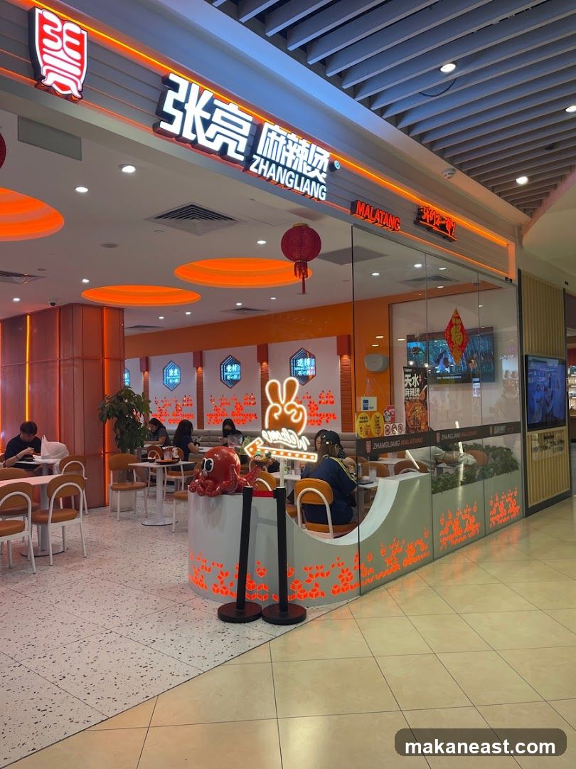The exterior of a Zhangliang Malatang restaurant with prominent signage. The restaurant features a modern interior with orange accents, tables, and chairs. Several patrons are seated inside. A decorative red octopus figure is placed near the entrance.