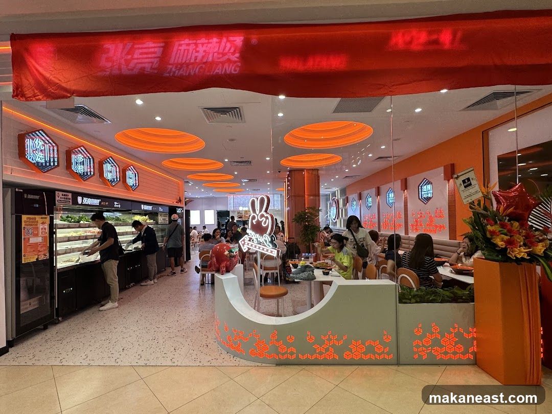 The exterior of a Zhang Liang Malatang restaurant. A red banner with the restaurant's name in Chinese and English hangs above the entrance. Inside, customers are seen selecting ingredients from a display case, and others are seated at tables. The interior is decorated with orange lighting and hexagonal wall art.