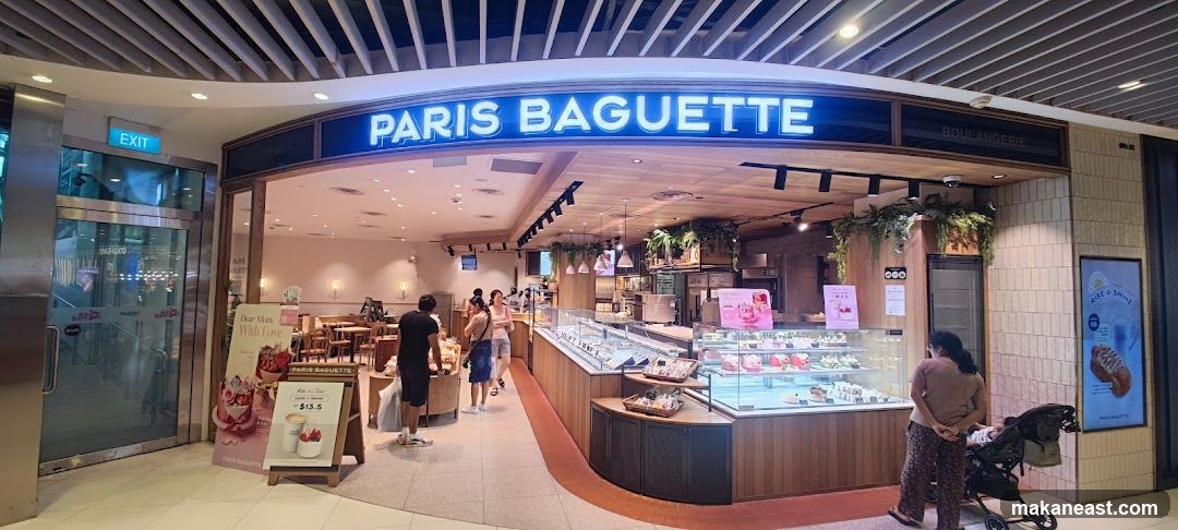 The exterior of a Paris Baguette bakery and cafe. The storefront features a large illuminated sign that reads "PARIS BAGUETTE" and "BOULANGERIE". Inside, display cases are filled with pastries and cakes. Seating areas with tables and chairs are visible. Several customers are browsing or waiting in line. An "EXIT" sign is visible on a glass door to the left. Promotional posters are displayed near the entrance.