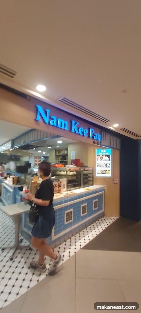 A woman walks past the storefront of Nam Kee Pau, a bakery. The shop has a blue and white tiled counter and a sign with the shop's name in blue neon letters. Behind the counter, baked goods are displayed in glass cases. A menu board with pictures of food items is visible on the right side of the storefront.