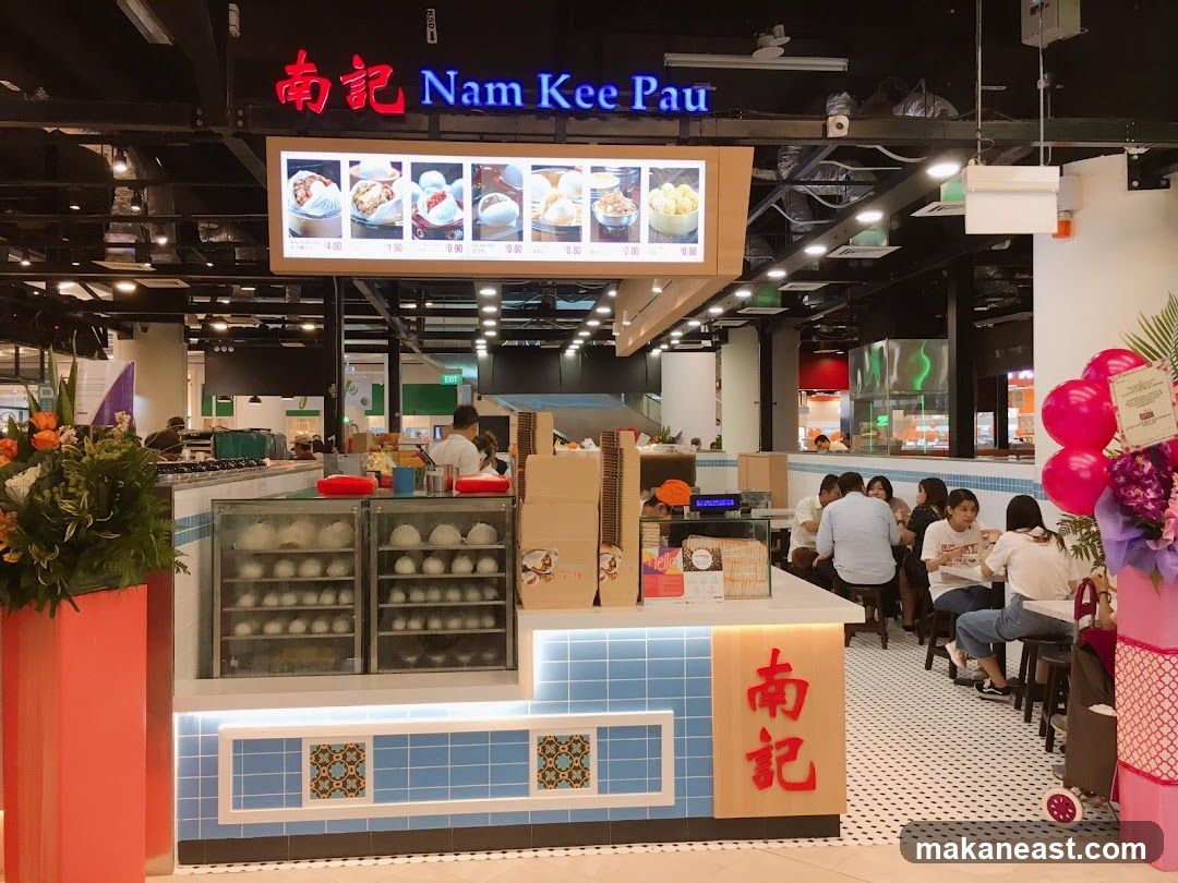 A food stall with a prominent sign that reads "Nam Kee Pau" in English and Chinese characters. Above the counter, a digital menu displays images of food items with prices. The counter itself has glass display cases filled with steamed buns. Customers are seated at tables in the background.