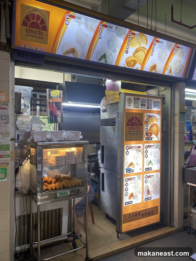 A food stall with a prominent illuminated sign displaying the business name "GENIE GO Gourmet on the Go" and images of various food items. Below the main sign, a smaller sign indicates the stall number "#01-157". To the left, a glass display case contains rows of packaged food items and fried snacks. To the right, a vertical menu board showcases different dim sum options with prices. The stall appears to be open for business.