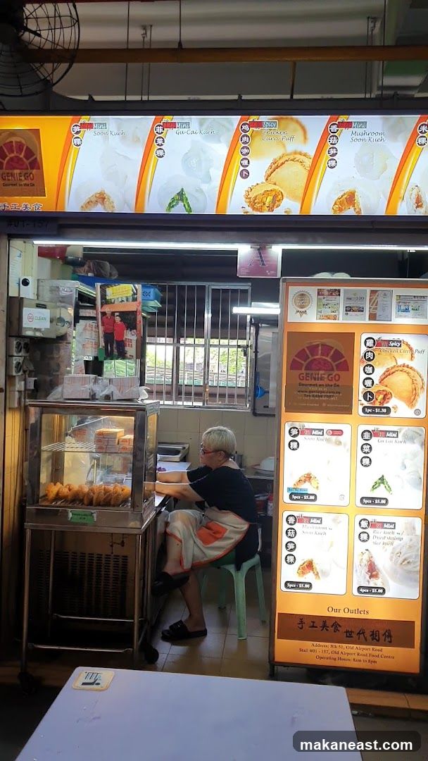 A food stall with a lit menu board displaying various food items. A person wearing an apron is seated behind a display case filled with fried food. The stall has a sign with the business name and logo.