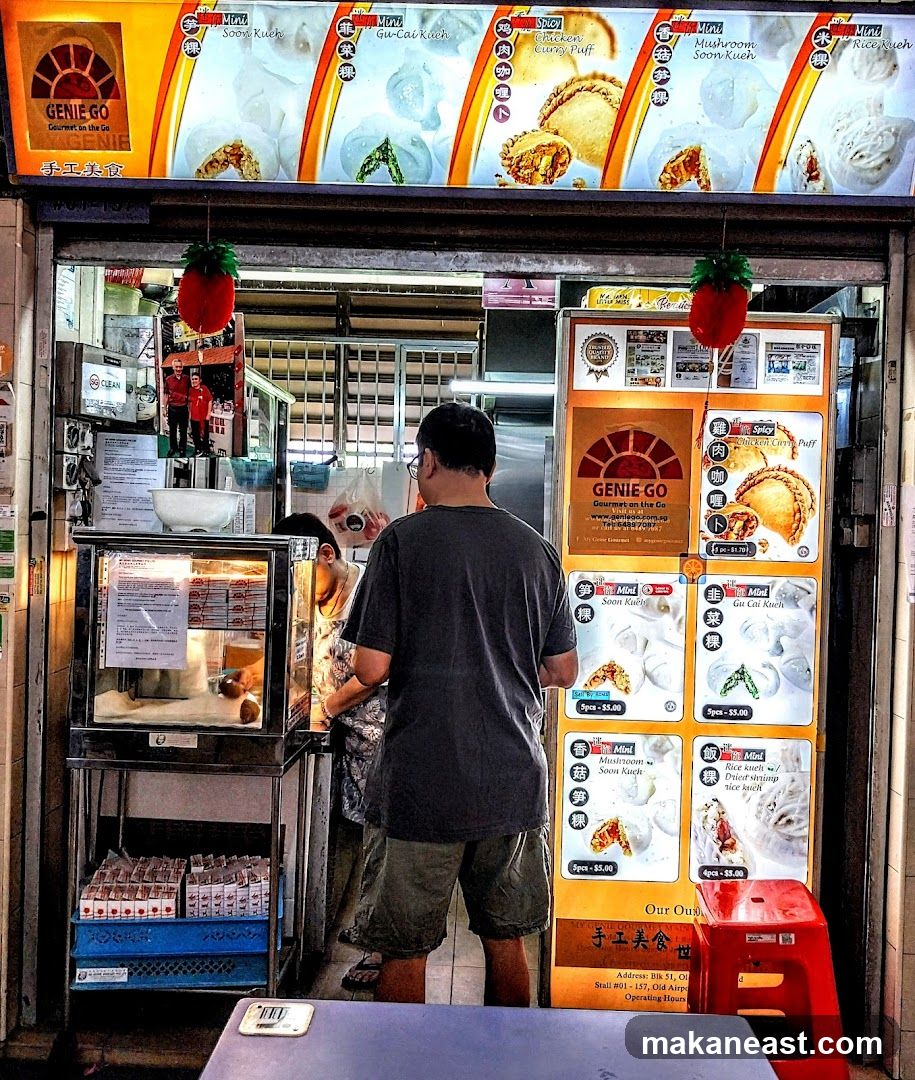 A food stall with a bright orange awning displaying menu items. A man is standing in front of the counter, facing away from the camera, looking at the menu. There are various food items displayed in a glass case and on posters.
