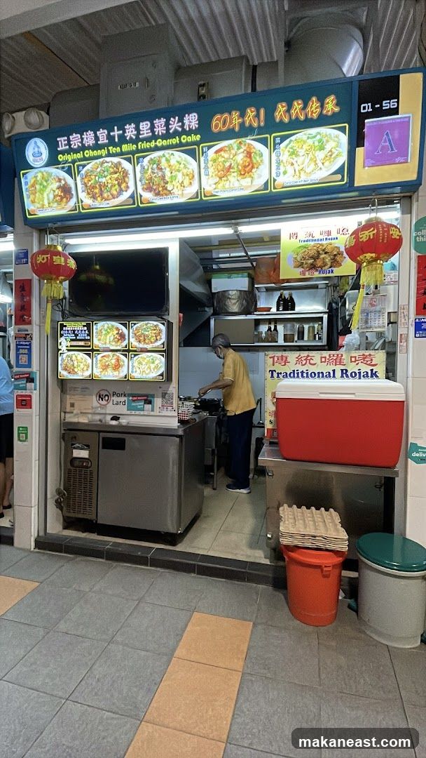 A food stall with a prominent sign displaying "Original Changi Ten Mile Fried-Carrot Cake" and images of the dishes. Below the main sign, there is a smaller sign for "Traditional Rojak". A person is seen cooking inside the stall. There are also refrigerators, a cooler, and various containers visible. A "NO Pork Lard" sign is also displayed.