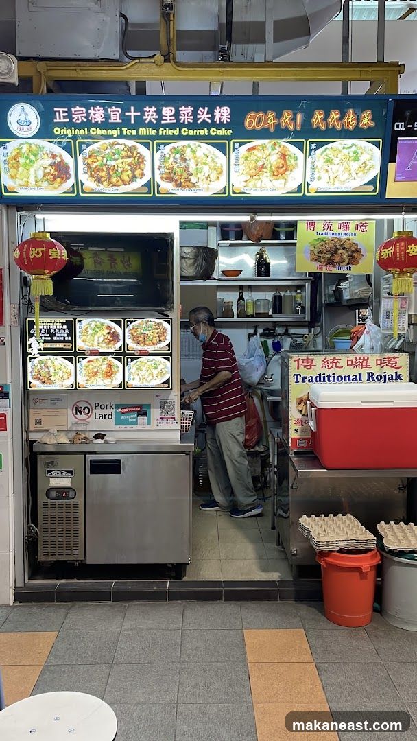 A food stall with a sign displaying "Original Changi Ten Mile Fried Carrot Cake" and images of various carrot cake dishes. Below the main sign, there are digital displays showing more food images and menu items. A "No Pork Lard" sign is also visible. A man in a striped shirt and mask is standing inside the stall. Another sign advertises "Traditional Rojak". Red lanterns hang on either side of the stall entrance.