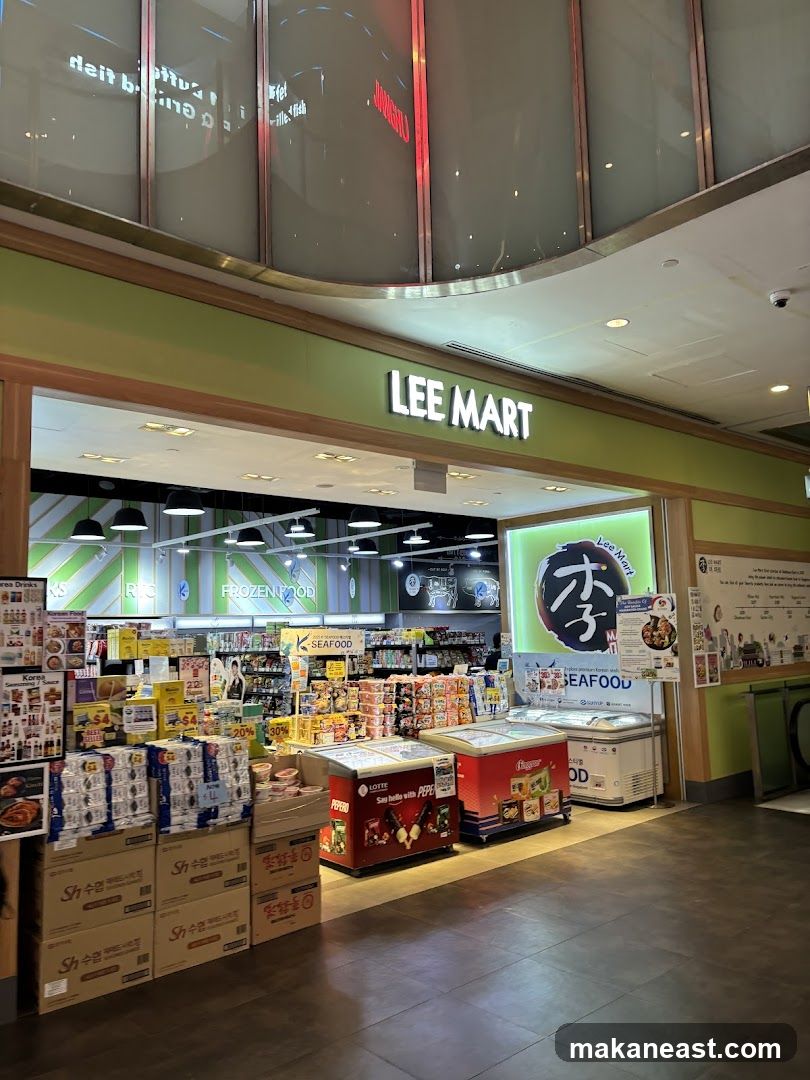The exterior of a store called Lee Mart, with its name prominently displayed above the entrance. The store appears to be a market selling Korean food and seafood, with various products visible inside, including frozen food and packaged goods. Refrigerated display cases are positioned outside the entrance.