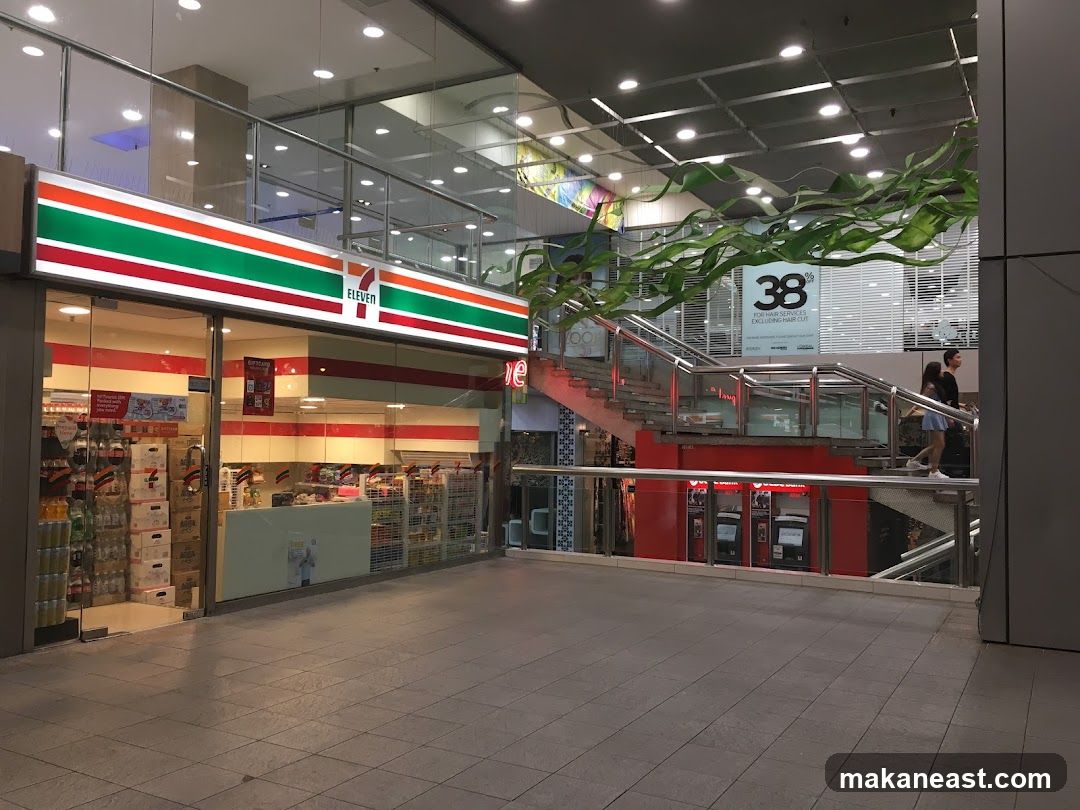 The exterior of a 7-Eleven convenience store with its distinctive red, green, and orange striped signage. The store entrance is visible, with shelves stocked with drinks and other products displayed behind the glass doors. To the right, a staircase leads up to another level of the building, with people walking up. A sign advertising "38% FOR HAIR SERVICES EXCLUDING HAIR CUT" is visible on the upper level.