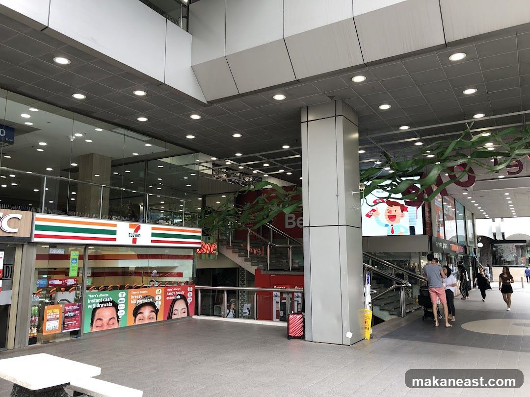 The exterior of a 7-Eleven convenience store with its prominent red, green, and white signage. The store has large glass windows displaying various promotional posters and products. To the right, a digital screen shows a cartoon character holding money. People are walking by on the sidewalk.