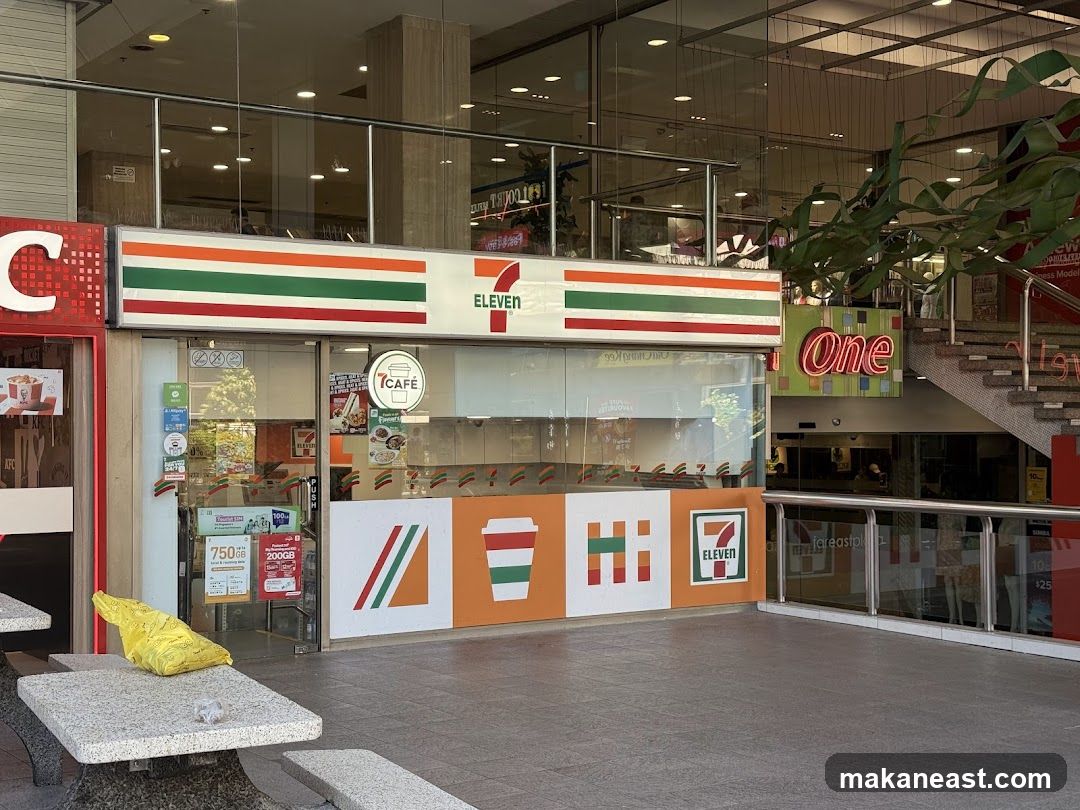 The exterior of a 7-Eleven convenience store is visible, featuring its signature red, green, and orange striped logo above the entrance. To the left, a KFC storefront is partially visible. The store has large glass windows displaying merchandise and a "7CAFÉ" sign. Decorative panels with geometric designs and the 7-Eleven logo are mounted on the lower exterior wall. A yellow plastic bag and some crumpled paper are on a nearby stone table.