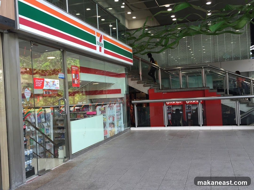 The exterior of a 7-Eleven convenience store with its distinctive red, green, and white striped signage. The entrance features glass doors and windows, revealing shelves stocked with various products inside. Adjacent to the store, a staircase leads up, with OCBC Bank ATMs visible below the stairs.
