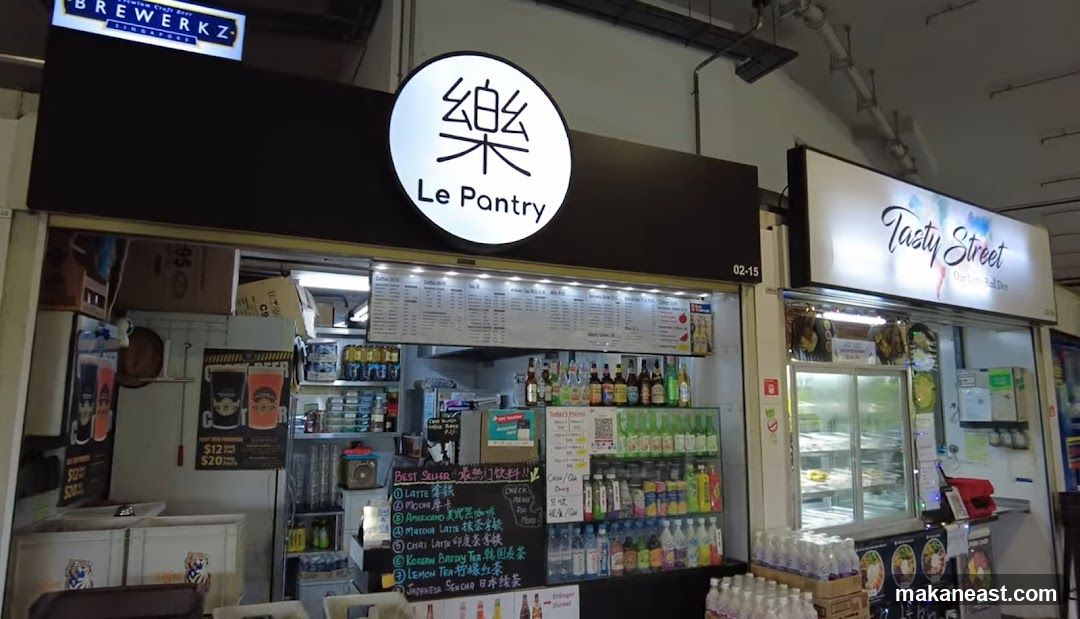A storefront for Le Pantry, a food and beverage stall. The stall has a large circular illuminated sign with the name "Le Pantry" and a Chinese character. Below the sign is a menu board displaying various drinks and food items. The stall is stocked with bottles of beverages and other products. To the right is another stall labeled "Tasty Street".