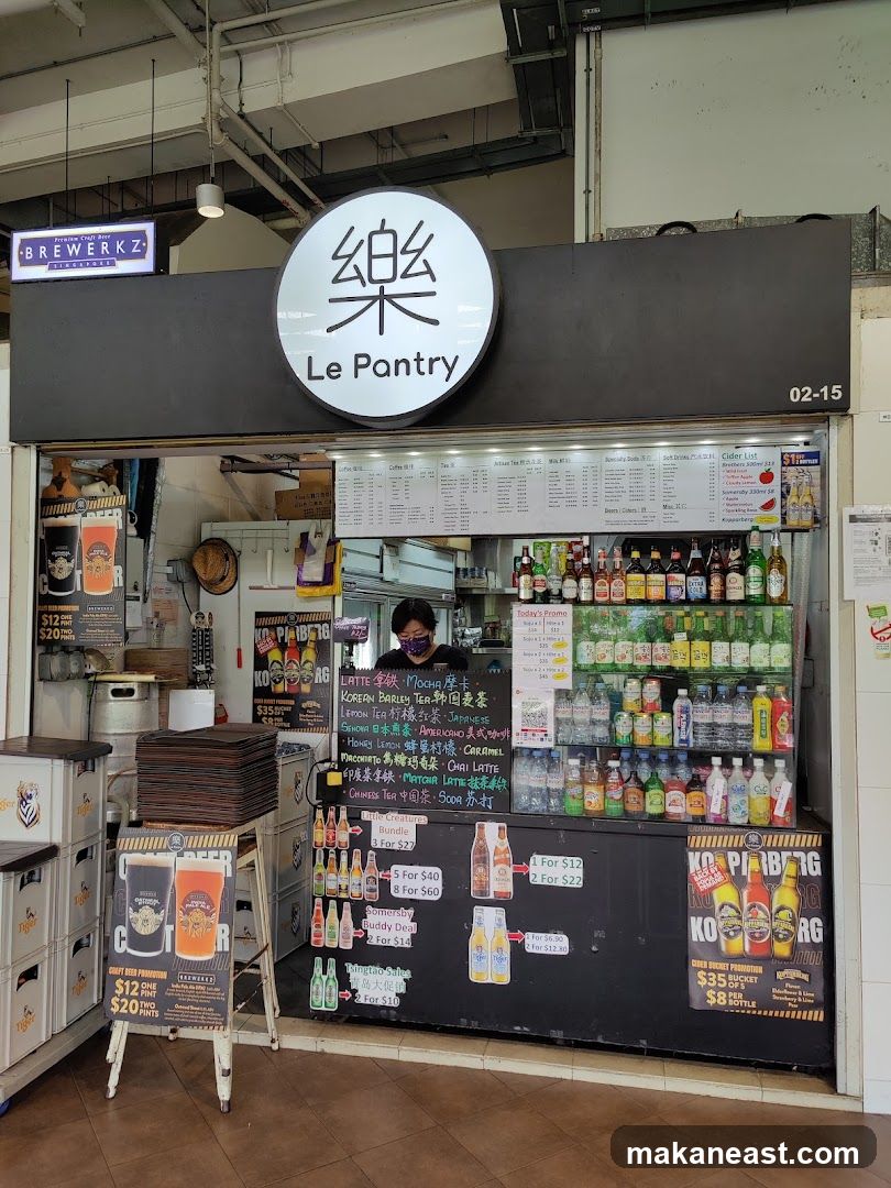 A shop front with a large circular sign displaying Chinese characters and the text "Le Pantry". Below the sign is a counter with various drinks and promotional materials. A person wearing a mask is visible behind the counter. There are also stacks of crates and promotional stands outside the shop.