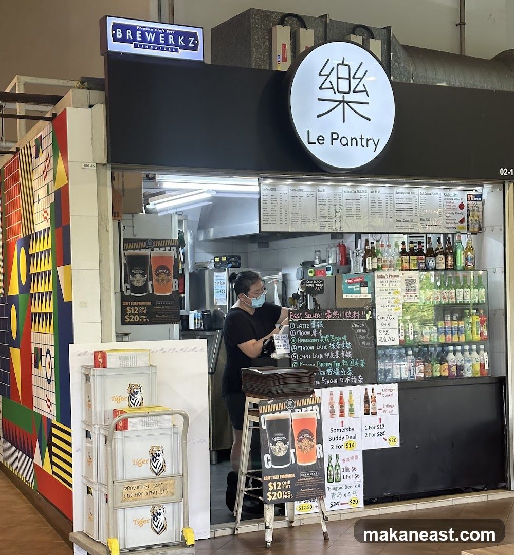 A food stall with a prominent circular sign featuring Chinese characters and the text "Le Pantry". Above the stall, a sign reads "BREWERKZ SINGAPORE". A person wearing a mask is standing behind the counter. Various drinks and promotional materials are displayed. Stacks of Tiger beer crates are visible to the left.