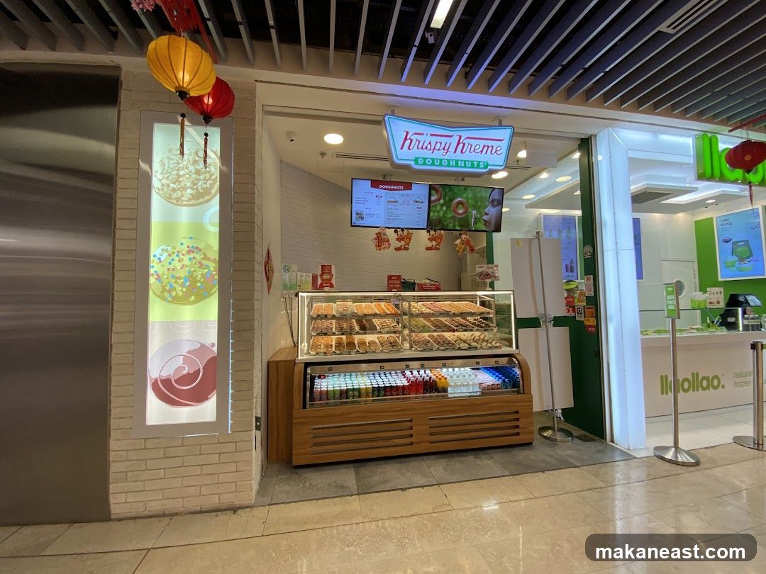 The exterior of a Krispy Kreme Doughnuts shop. A display case filled with various doughnuts is visible, along with a refrigerated section below containing drinks. Above the display, digital screens show menu items and promotional images. To the right, a Yoogoo shop is also visible. Decorative lanterns hang from the ceiling.