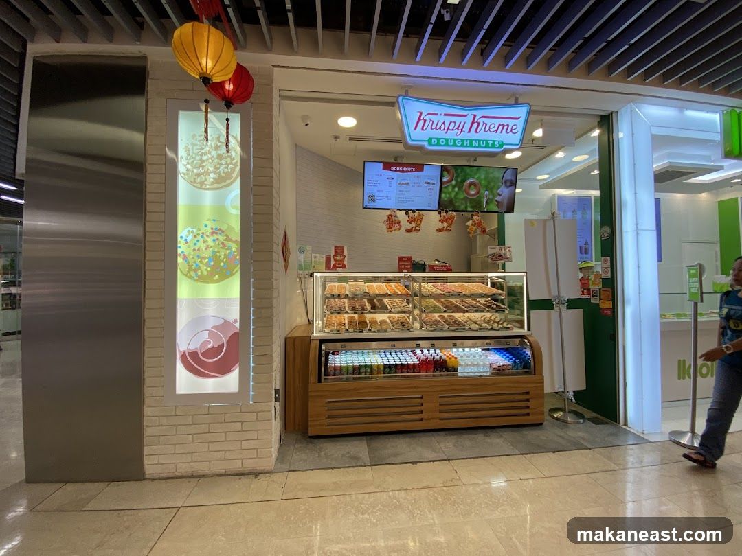A Krispy Kreme Doughnuts shop front with a display case full of doughnuts and a refrigerated section with drinks. There are also decorative elements on the left side of the shop, including images of doughnuts and lanterns hanging above. A digital menu board is visible above the display case.