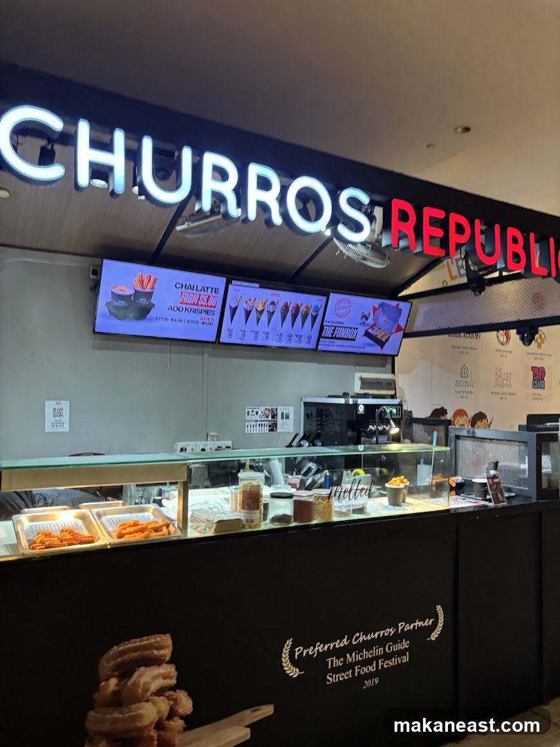 A brightly lit storefront with a large neon sign reading "CHURROS REPUBLIC". Below the sign, digital menu boards display various offerings, including "CHAI LATTE" and "THE FUNBOX". The counter area features display trays with churros and other food items, along with a coffee machine. The front of the counter has a graphic of churros and text stating "Preferred Churros Partner The Michelin Guide Street Food Festival 2019".