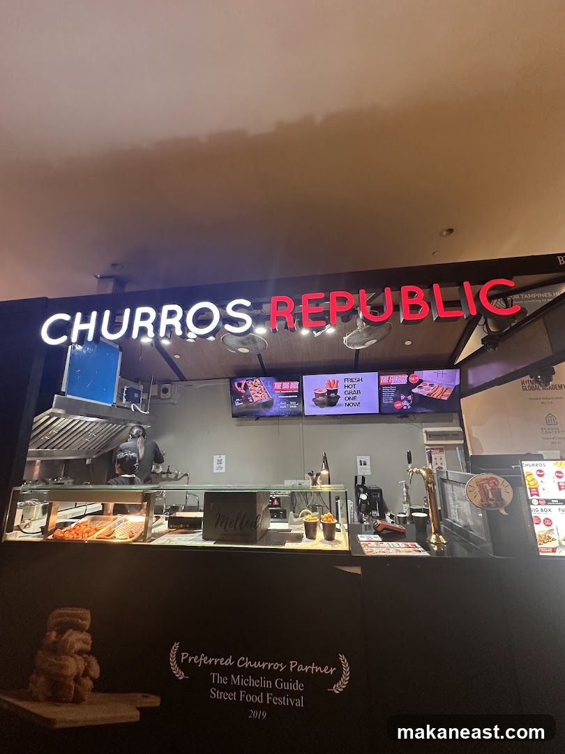 A brightly lit storefront for Churros Republic, featuring a large neon sign. The counter displays various churro preparations and toppings. Digital screens above the counter show promotional images of churros. A sign on the lower part of the counter indicates "Preferred Churros Partner" for the Michelin Guide Street Food Festival 2019.
