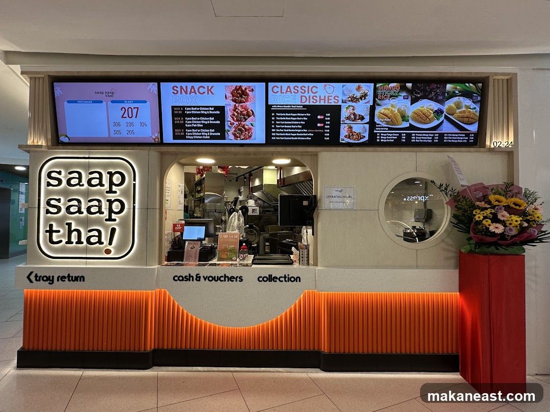A food stall with a prominent "saap saap tha!" logo. Digital screens display order numbers and menu items. The counter area shows a cash and vouchers collection point.