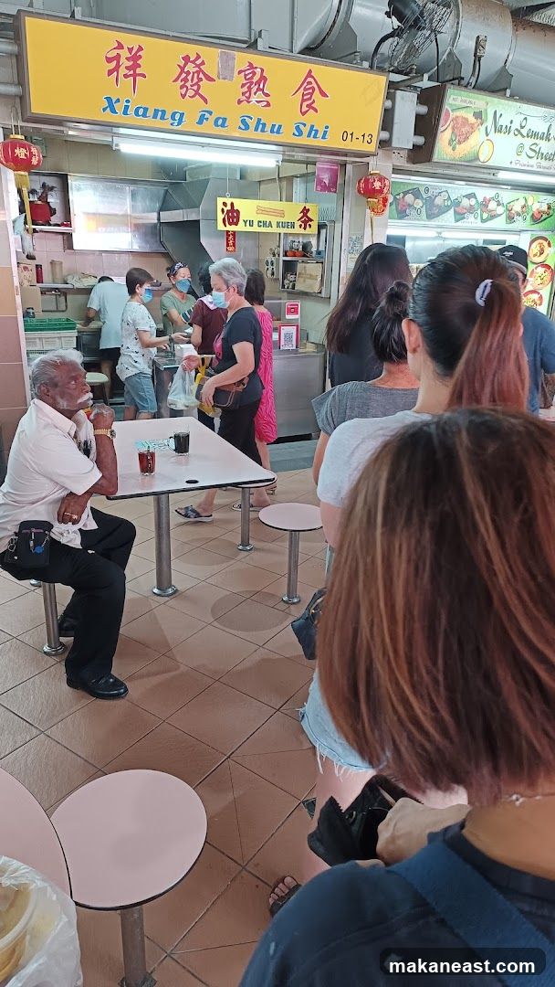 A yellow sign with Chinese characters and "Xiang Fa Shu Shi" written in blue is visible above a food stall. Below the sign, a smaller yellow sign reads "YU CHA KUEN" with Chinese characters. People are queuing at the stall, and one man is seated at a table. Other food stalls with menus are visible to the right.