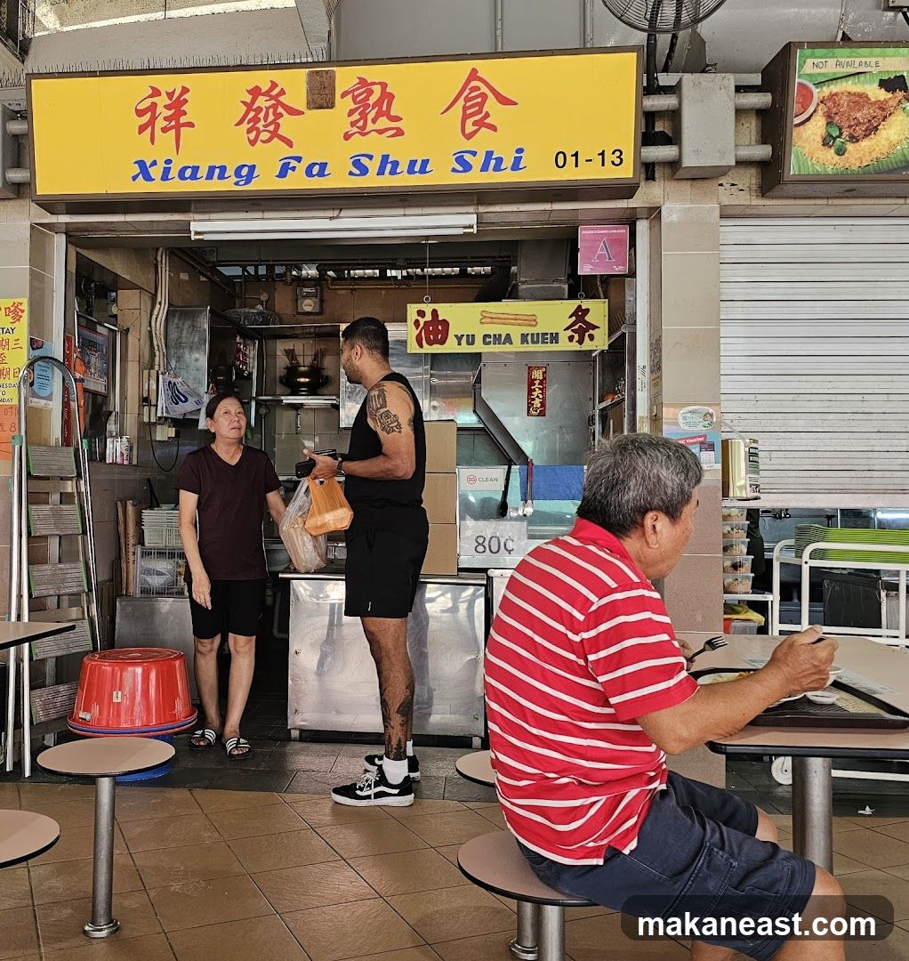 A food stall with a yellow sign that reads "Xiang Fa Shu Shi" in both Chinese and English. Below the sign, there are various food items and people interacting. One person is ordering food, while another is seated at a table eating. The stall appears to be in an outdoor food court or hawker center.