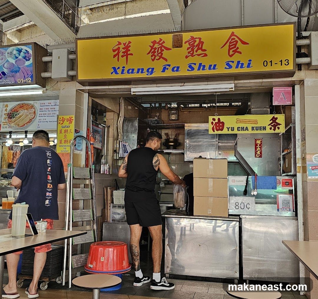 A food stall with a yellow sign that reads "Xiang Fa Shu Shi" in Chinese and English. Two men are visible, one ordering and the other waiting. There are boxes stacked on the counter and a sign advertising "YU CHA KUEH" for 80 cents.