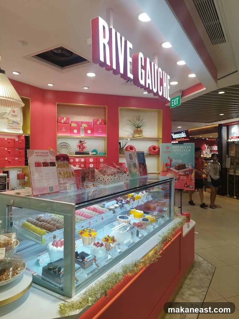 A brightly lit bakery with a pink interior and a display case filled with colorful pastries and cakes. The RIVE GAUCHE logo is prominently displayed above the counter.