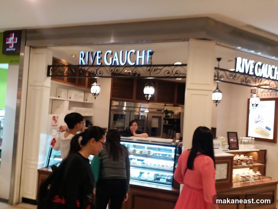 The exterior of a patisserie with the name "RIVE GAUCHE" displayed prominently above the entrance. Inside, display cases show various pastries and cakes. Several people are visible inside and near the entrance.