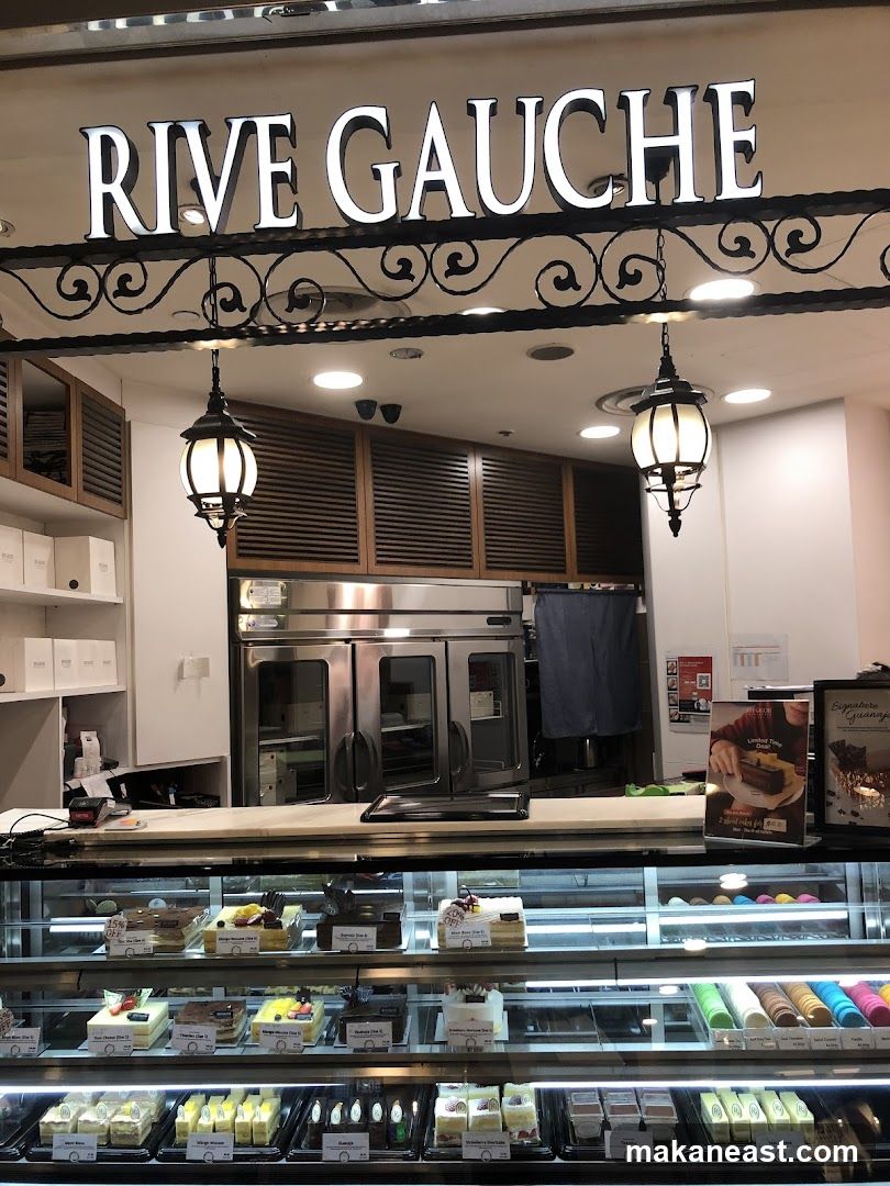 The exterior of a bakery with a large illuminated sign that reads "RIVE GAUCHE". Below the sign, there are two hanging lanterns. A display case filled with various cakes and pastries is visible. Behind the counter, there are stainless steel ovens and shelving units.