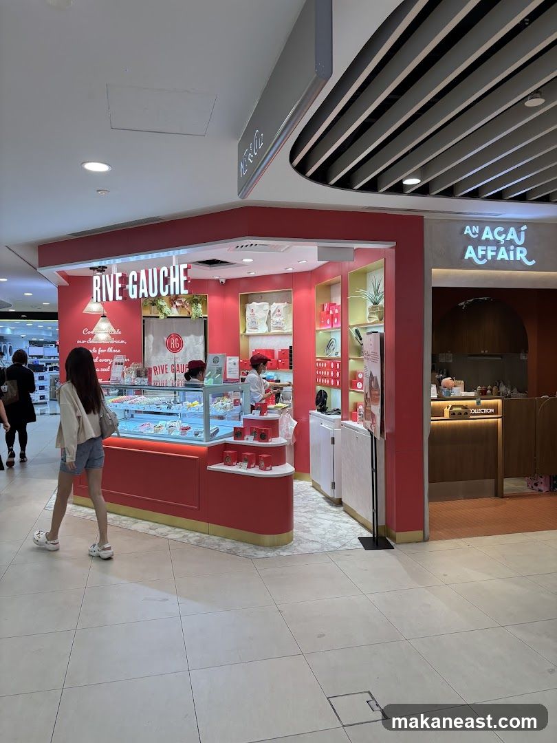 A bright pink and red storefront for Rive Gauche, a patisserie, is visible in a mall. The shop features a display case filled with colorful pastries and cakes. Behind the counter, staff members in red uniforms are visible. The shop's name, RIVE GAUCHE, is prominently displayed in large white letters above the entrance. To the right of Rive Gauche, another shop with the name AN ACAI AFFAIR is visible.