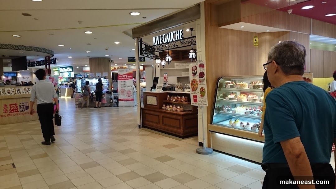 The exterior of a bakery shop called Rive Gauche, located in a mall. The shop has a display case filled with various cakes and pastries. Above the counter, there is a sign with the shop's name and decorative lamps. There are also posters displaying cakes.
