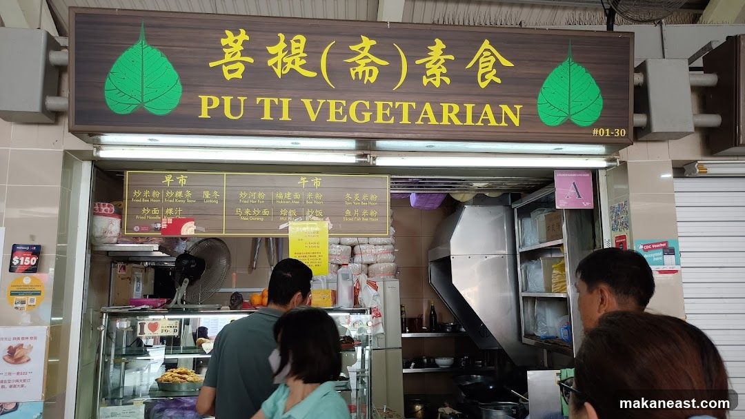 A vegetarian food stall with a wooden sign displaying the name "PU TI VEGETARIAN" in yellow Chinese characters and English. Below the sign is a menu board with two sections, "早市" (Morning Market) and "午市" (Afternoon Market), listing various noodle and rice dishes. People are seen queuing and ordering food at the counter.