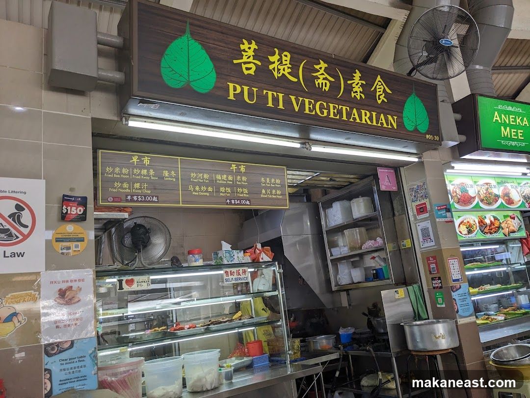 A vegetarian food stall with a prominent sign displaying "PU TI VEGETARIAN" in English and Chinese. Below the main sign, a menu board lists various dishes categorized into "早市" (Morning Market) and "午市" (Afternoon Market). The stall has a display counter with food items and a self-service area. To the right, another stall labeled "ANEKA MEE" is visible.