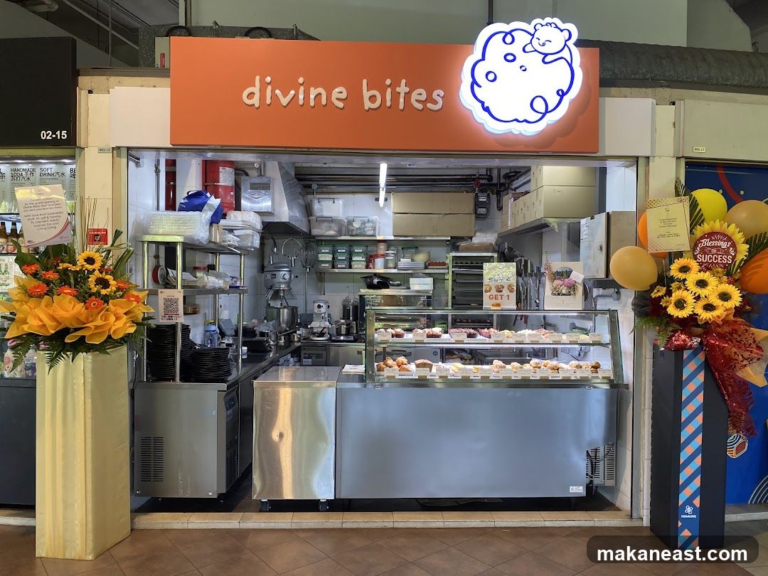 The exterior of a bakery with a bright orange sign that says "divine bites" and a cartoon bear logo. In the display case, there are many cupcakes and pastries. There are also floral arrangements and balloons outside the shop.