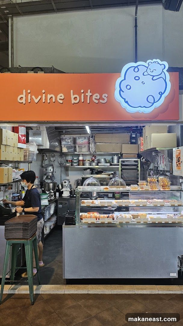A bakery storefront with the name "divine bites" displayed prominently on an orange sign above the entrance. A cartoon character is depicted on a cloud next to the text. Inside, a display case showcases various pastries and baked goods. A staff member wearing a mask is visible behind the counter, interacting with a register. Shelves stocked with ingredients and baking equipment are also visible in the background.