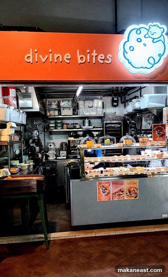 The exterior of a bakery with an orange awning displaying the name "divine bites" and a cartoon cloud logo. Inside, a display case shows various pastries and cakes. Shelves behind the counter are stocked with baking supplies and equipment.