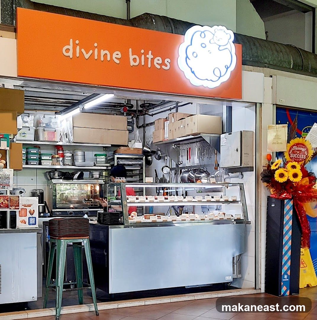 A bakery storefront with an orange sign that says "divine bites" and a cartoon cloud logo. Inside, there is a display case filled with pastries, shelves stocked with ingredients and equipment, and a person working behind the counter.