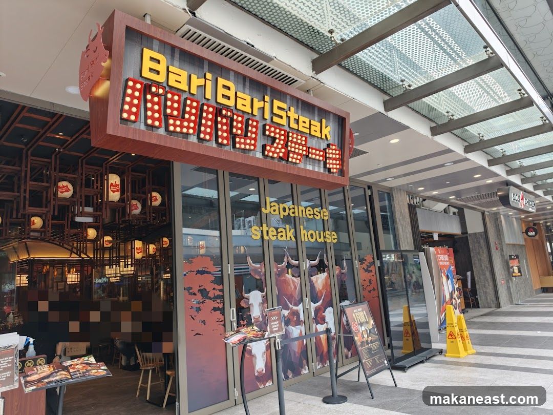 The exterior of a Japanese steak house with a prominent sign that reads "BariBari Steak" in large yellow letters with red accents. Below the main sign, it says "Japanese Steak house" in yellow text. The entrance features large glass doors and windows decorated with images of cattle. Inside, traditional Japanese lanterns are visible. A menu stand is placed outside the entrance.
