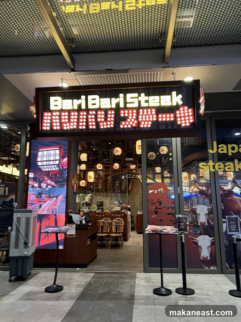 The exterior of a restaurant called Bari Bari Steak. The sign above the entrance is illuminated with red and yellow lights. Through the glass doors, tables and chairs are visible, along with some decorative lanterns. To the right of the entrance, a large graphic of cows is displayed on the glass.
