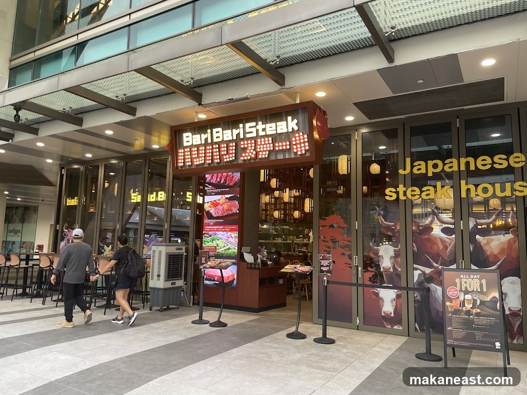 The exterior of a Japanese steakhouse with a prominent neon sign that reads "Bari Bari Steak". The storefront features large glass doors and windows decorated with images of cows and text that says "Japanese steak house". There are also promotional displays and a queue barrier outside.