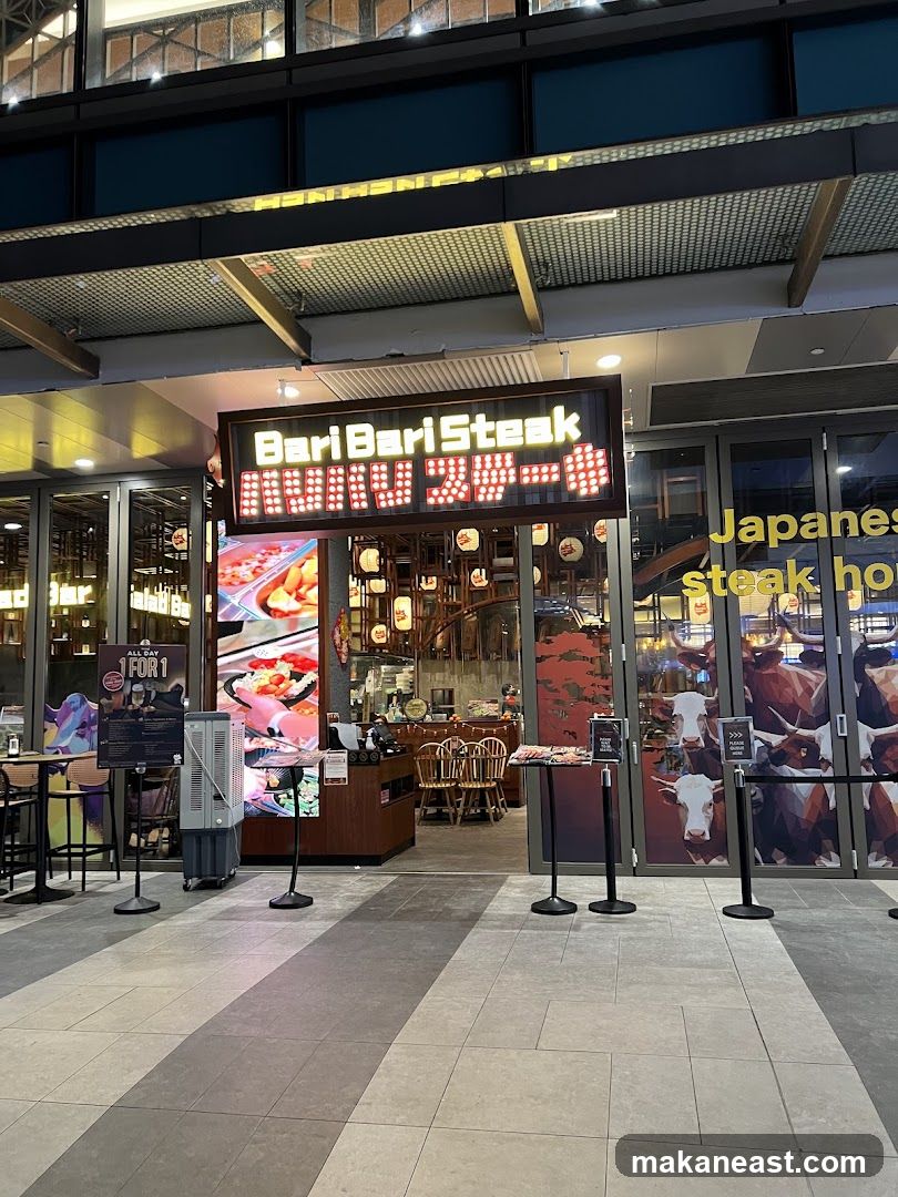 The exterior of a restaurant called Bari Bari Steak. A large illuminated sign above the entrance reads "BariBari Steak" in English and Japanese characters. To the right, a glass door has text that says "Japanese steak house". The entrance area shows tables and chairs, and a display of food. Stylized images of cows are visible on the glass doors to the right.
