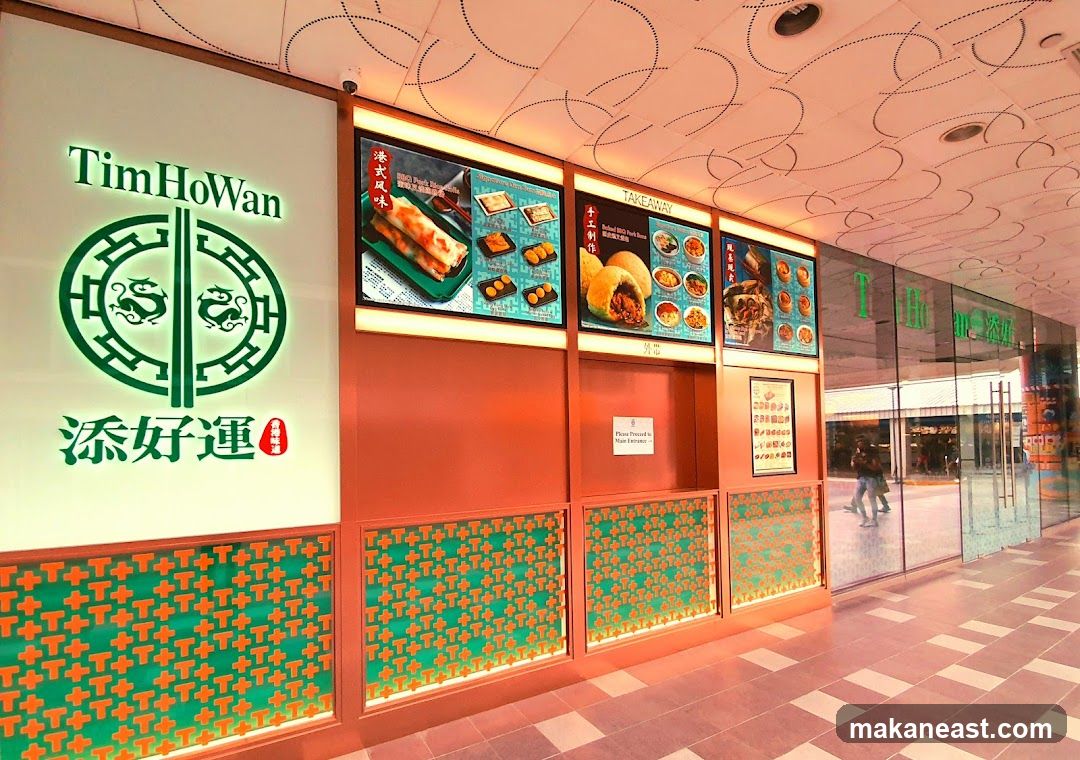 The exterior of a Tim Ho Wan restaurant with its logo and menu displays. The facade features a large illuminated sign with the restaurant's name and logo, alongside digital menu boards showcasing food items. The lower section of the facade is decorated with a repeating pattern. The entrance to the restaurant is visible through glass doors.