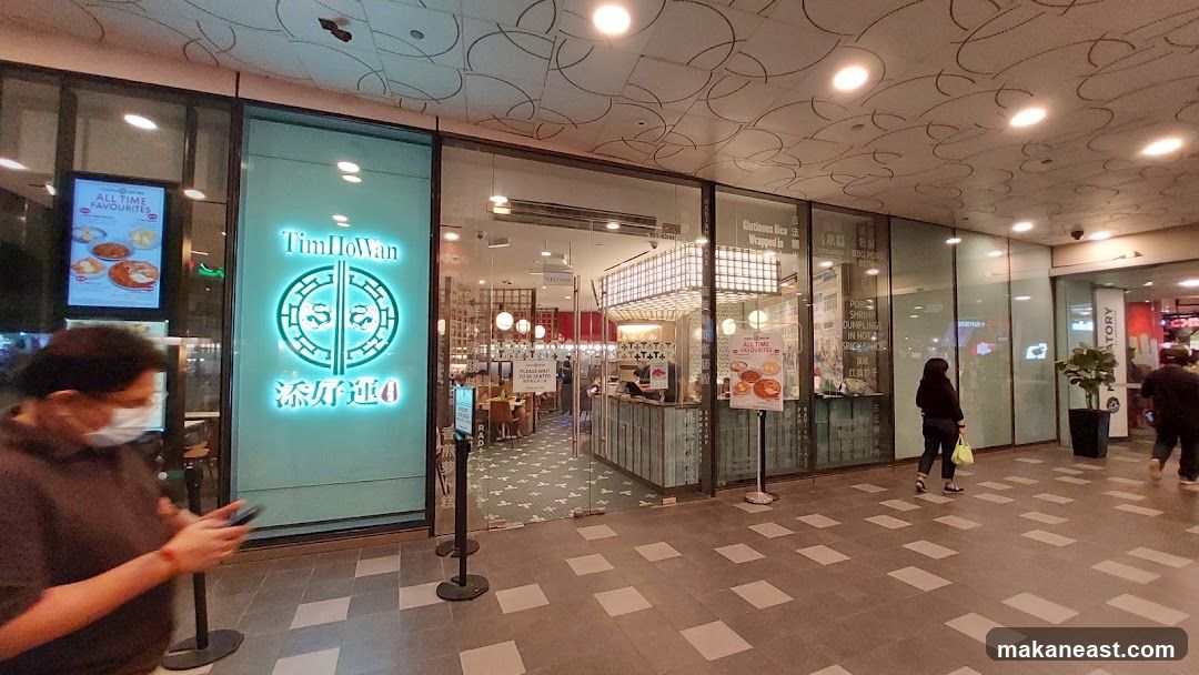 The exterior of a Tim Ho Wan restaurant with large glass windows and doors. A prominent neon sign with the restaurant's logo and name is visible on the left side of the entrance. Inside, tables and chairs are set up, and a counter area is visible. A digital display showing "ALL TIME FAVOURITES" with pictures of food is on the left. Another display with similar content is on the right.