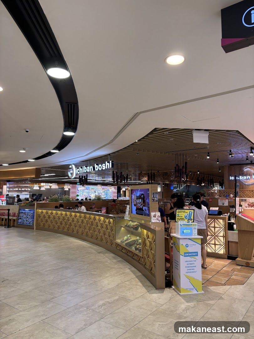 The exterior of a restaurant with the name "Ichiban Boshi" visible on the signage above the entrance. The restaurant has a modern design with a curved counter displaying food items. People are seen dining inside and staff are visible behind the counter.
