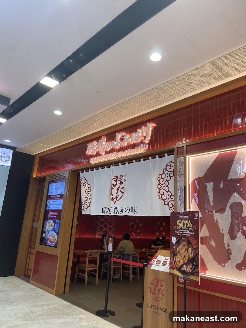 The exterior of a Japanese restaurant called Butahage. The entrance features a red tiled facade with a large white noren curtain displaying Japanese characters and a logo. Above the curtain, a neon sign spells out the restaurant's name in Japanese. To the left of the entrance, a digital display shows menu items and a QR code. Inside, wooden tables and chairs are visible, with patrons seated. A promotional poster for a 50% discount on Tuesdays is displayed near the entrance.