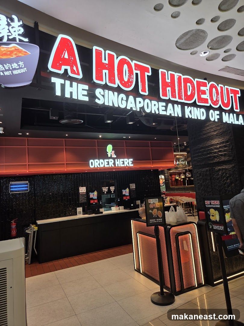 The exterior of a restaurant called "A Hot Hideout" with prominent red and white signage. Below the main sign, it reads "THE SINGAPOREAN KIND OF MALA". An "ORDER HERE" sign is visible above the counter area. The entrance features a modern design with illuminated panels.