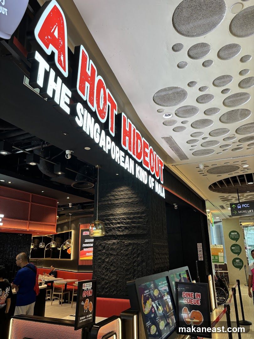 The exterior of a restaurant with a large illuminated sign that reads "A HOT HIDEOUT THE SINGAPOREAN KIND OF MALA". The entrance is visible with people inside. There are also digital menu boards outside the entrance.