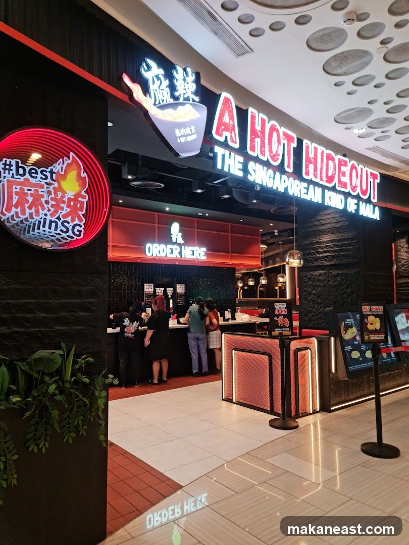 The exterior of a restaurant with prominent signage that reads "A HOT HIDEOUT THE SINGAPOREAN KIND OF MALA". To the left, a circular neon sign displays "#best 麻辣 in SG". An "ORDER HERE" sign is visible above the counter area where customers are queuing.