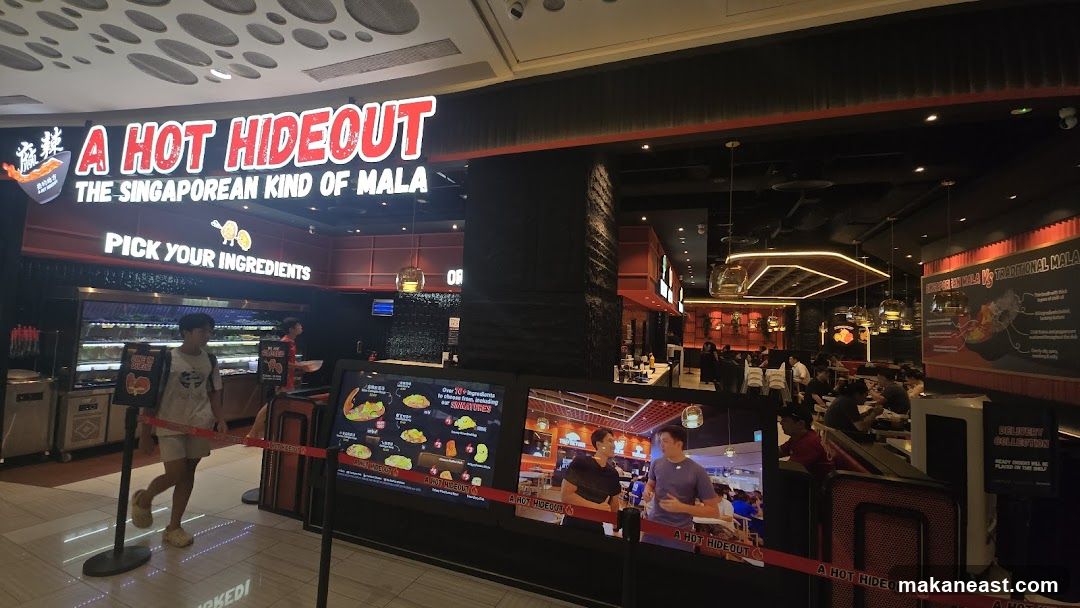 The exterior of a restaurant called A Hot Hideout, which specializes in Singaporean Mala. The signage is prominent, with the name "A HOT HIDEOUT" in large red letters above the tagline "THE SINGAPOREAN KIND OF MALA". Below this, it says "PICK YOUR INGREDIENTS". The entrance area features displays of ingredients and digital screens showing food items and promotions.